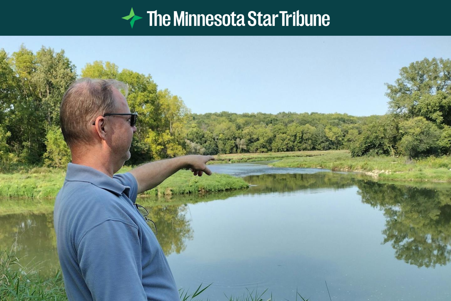 A man stands alongside a river and points to the Root River restoration project site.